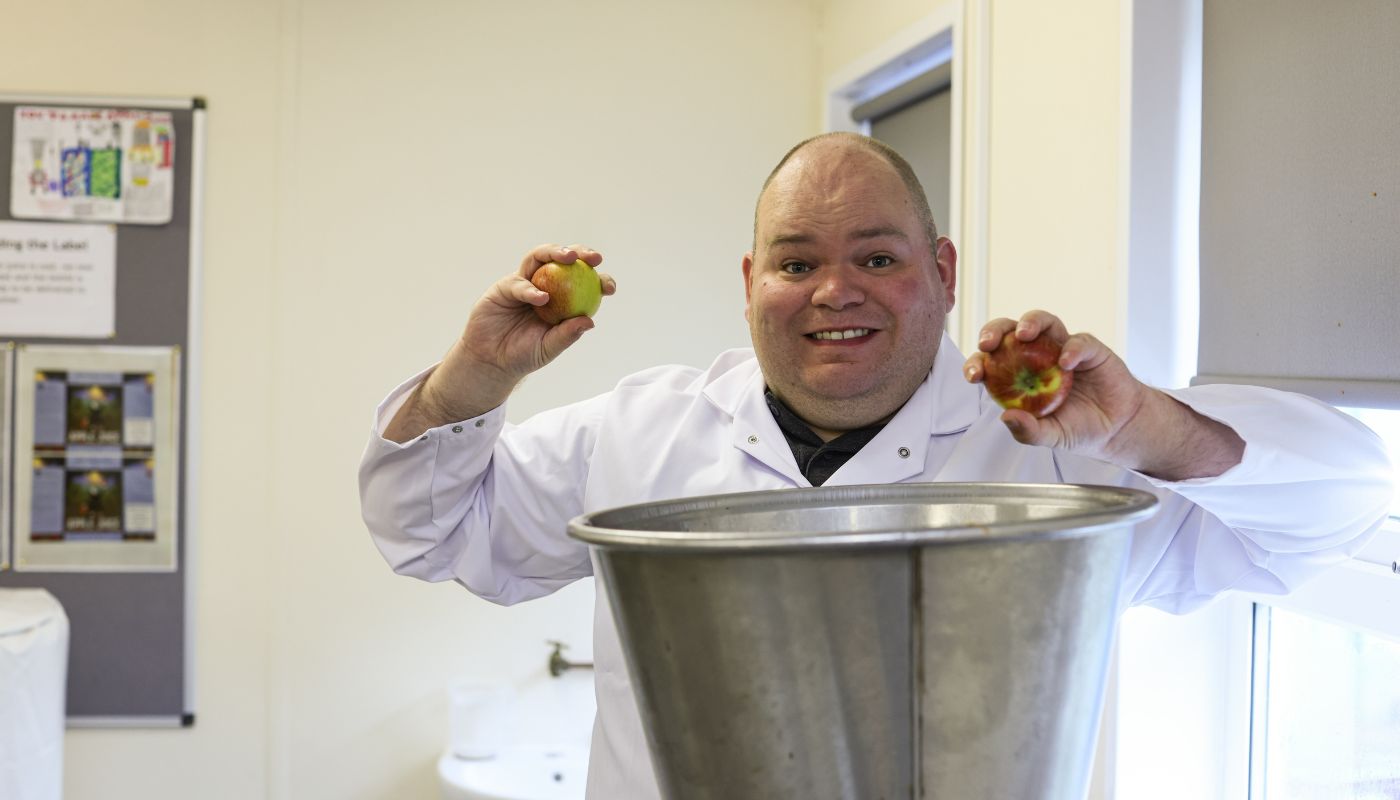 A man in a white coat smiles and holds up two apples in-front of a fruit mill for making apple juice.